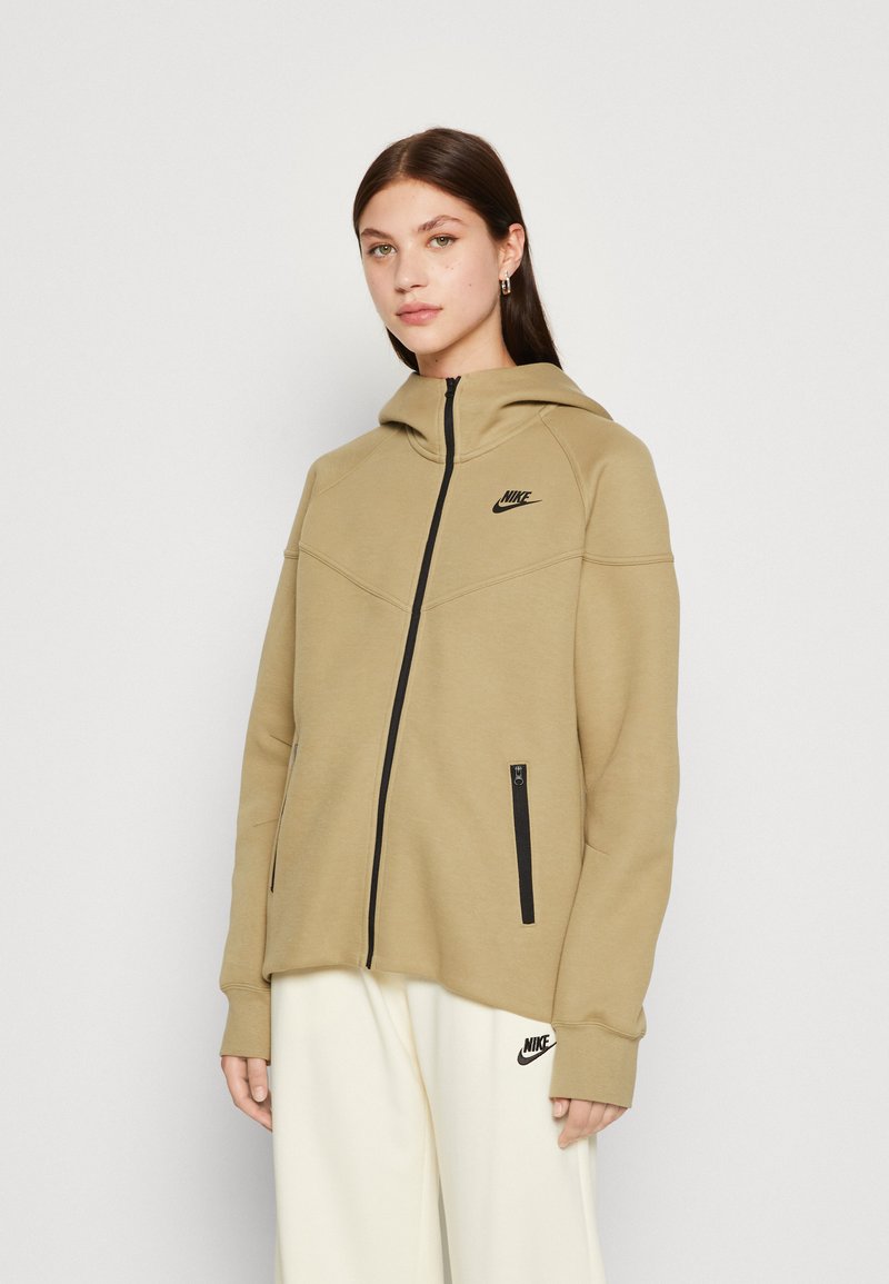 Beige krēmo krāsā Nike kapuce ar rāvējslēdzēju, melnu logotipu un divām melnām rāvējslēdzēja kabatām. Gluda tekstūra un sportisks piegriezums.