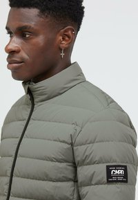 Green puffer jacket with horizontal quilted design, high collar, and zip closure. Features a branded patch on left sleeve.
