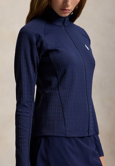 Γυναίκα που φοράει μπλε σκούρο μπουφάν με φερμουάρ και σχέδιο πλεκτής καλώδιο καθώς και μικρό λευκό λογότυπο στο στήθος.