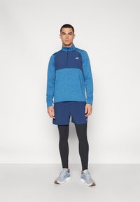 Maglietta sportiva blu a maniche lunghe con zip a quarti, abbinata a pantaloni corti blu e leggings neri. Branding notevole sul petto. Scarpe da corsa grigie.