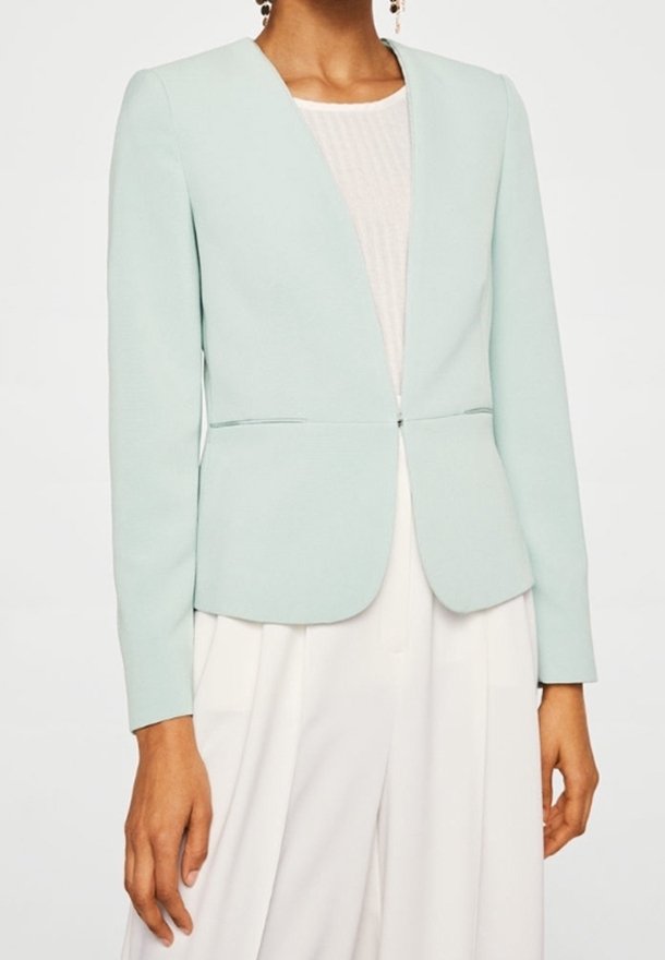 Femme portant un blazer bleu clair sur un haut blanc et un pantalon blanc taille haute, avec des manches longues et une fermeture à l'avant unique.