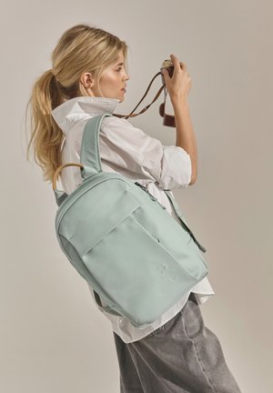 Blonde vrouw in wit shirt en grijze broek, die een vintage camera vasthoudt en een lichtgroene rugzak met houten handvatdetail draagt.