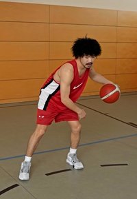 Rød basketballtrøye med svart og hvite detaljer, grå sneakers og en basketball i hånden. Står på et gymgulv, fokusert og i bevegelse.