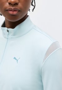 Άτομο που φοράει αθλητικό τοπ με φερμουάρ σε ανοιχτό γαλάζιο χρώμα, με λογότυπο Puma και διαπνέον σχέδιο στο ύψος του ώμου, εμφανιζόμενο από το πηγούνι μέχρι το ανώτερο μέρος του κορμού.