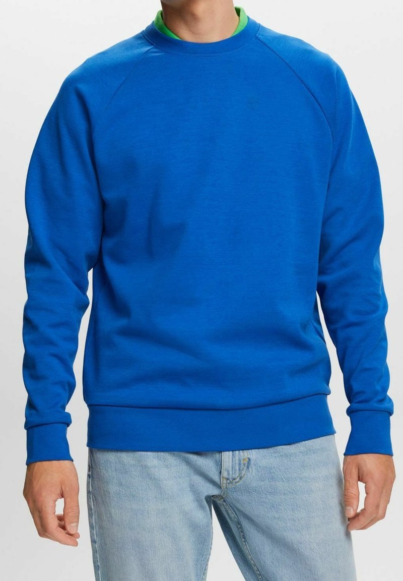 Un homme porte un sweat-shirt bleu vif à col rond par-dessus une chemise verte à col et un jean bleu clair, debout devant un fond blanc uni.