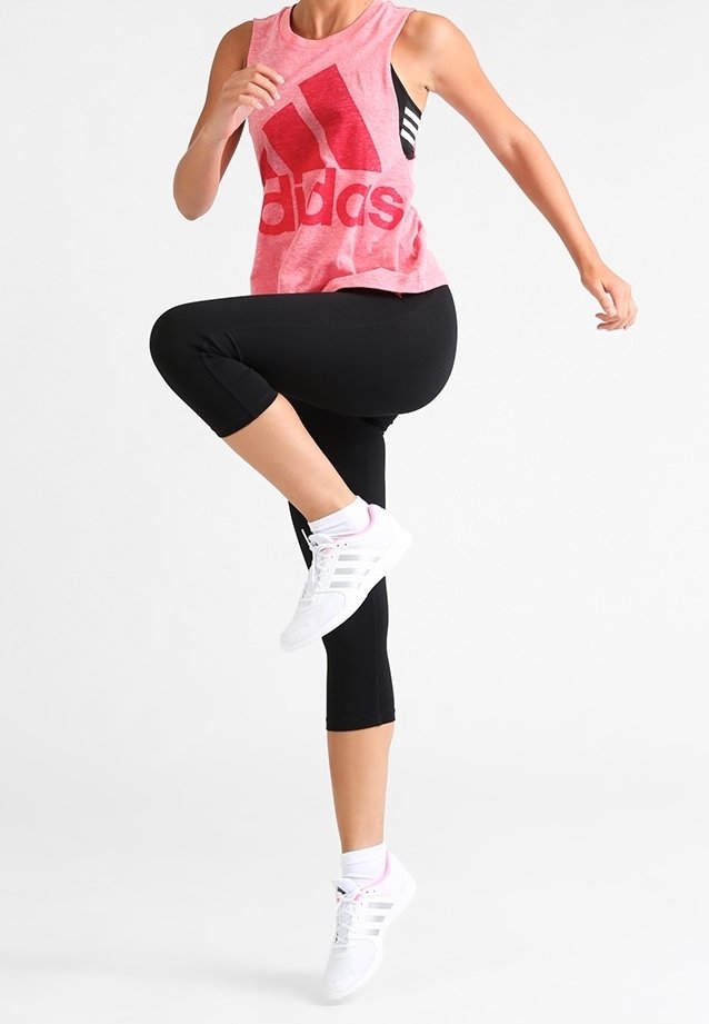 Femme en leggings noirs et débardeur rose Adidas levant un genou en plein exercice, portant des baskets blanches Adidas sur fond blanc.