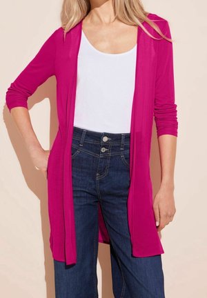 Kvinde iført en lys pink lang cardigan over en hvid top med rund hals og mørkeblå højtaljede jeans.