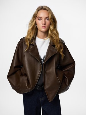 Braune Lederbikerjacke mit Reverskragen, Reißverschlussverschlüssen und übergroßem Schnitt. Getragen über einem Grafik-T-Shirt und dunklen Jeans.