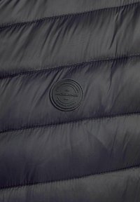 Schwarze Pufferjacke aus Material mit genähten Linien, einem runden, geprägten Logo-Patch mit dem Schriftzug "JACK & JONES" und einer glatten Textur.