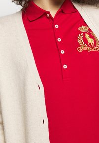 Polo en coton rouge avec col et fermeture à trois boutons, arborant un logo brodé doré. Gilet en tricot beige avec ouverture sur le devant et fentes sur les côtés.