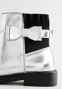 Bota de tobillo metálica plateada con un panel elástico negro, rematada con un lazo decorativo y un bajo tacón de goma negro. Material sintético texturizado.