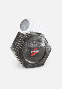 G-SHOCK-Uhr in einer schwarzen hexagonalen Dose. Die Uhr hat ein weißes Armband und ein rundes Ziffernblatt mit schwarzer Anzeige. Die Dose verfügt über ein erhabenes Logo und eine strukturierte Oberfläche.