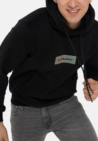 Haut à capuche noir avec une poche kangourou à l'avant, arborant un patch logo en blocs de couleur beige et vert. Jean gris avec effet délavé.