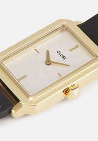 Montre rectangulaire dorée avec un cadran blanc, des aiguilles dorées et un bracelet en cuir noir. Présente un design minimaliste et un détail de couronne hexagonale.