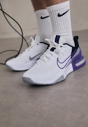 Baskets Nike blanches et violettes portées avec des chaussettes blanches Nike sur un sol gris, à côté d’un câble noir.