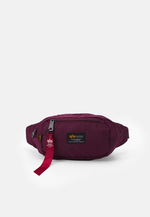 Bordeauxfarbene Bauchtasche mit vorderer Reißverschlusstasche, schwarz-gelbem Logo-Patch und rotem Stoffetikett am Reißverschluss.
