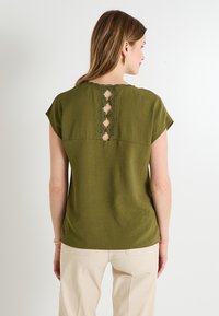 Haut olive vert à manches courtes avec un dos texturé présentant un panneau en dentelle et des découpes. Tissu lisse, coupe décontractée.