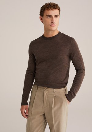 Hombre con suéter de manga larga marrón y pantalones beige con pliegues, posando con una mano en el bolsillo contra un fondo liso.