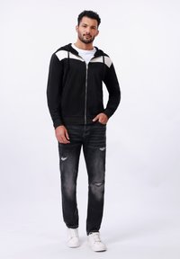 Sweat à capuche noir zippé avec des accents blancs sur les épaules, associé à un jean noir déchiré et des baskets blanches. Tissu lisse, coupe décontractée.