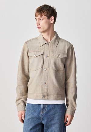 Junger Mann trägt eine beige Knopfleisten-Denimjacke über einem weißen Hemd und blauen Jeans, blickt nach links vor schlichtem Hintergrund.