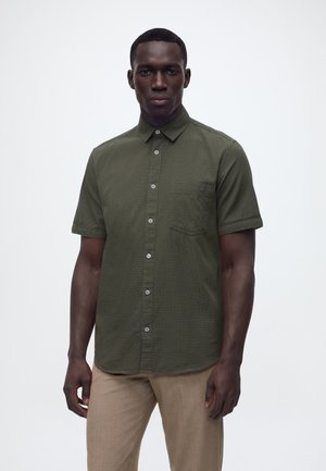 Άνδρας που φοράει πουκάμισο με κοντά μανίκια σε ελιά πράσινο χρώμα με διακριτικό σχέδιο και μπεζ παντελόνι, στέκεται μπροστά σε απλό φόντο.