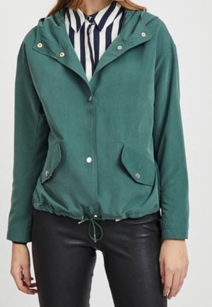 Mujer con chaqueta con capucha de color verde azulado y bolsillos frontales, sobre una camiseta a rayas blancas y negras y pantalones de cuero negro.