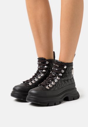 Černé kotníkové boty vyrobené z prošívaného materiálu, s metalickými očky, robustními černými podrážkami a kontrastními šňůrkami s texturovanými akcenty.