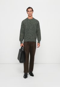 Man wearing a green knit sweater, brown pants, and black boots, holding a black leather tote bag against a white background.