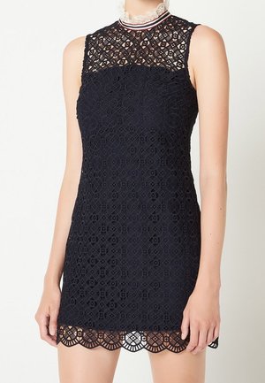 Czarna sukienka bez rękawów z geometrycznymi wzorami, z wysokim kołnierzem ozdobionym białą koronką i pasami, z falowanym dołem powyżej kolana.