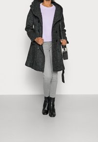 Cappotto grigio in misto lana con cappuccio, bottoni doppi, e cintura, abbinato a un maglione lilla e jeans grigi, stivali neri alla caviglia.