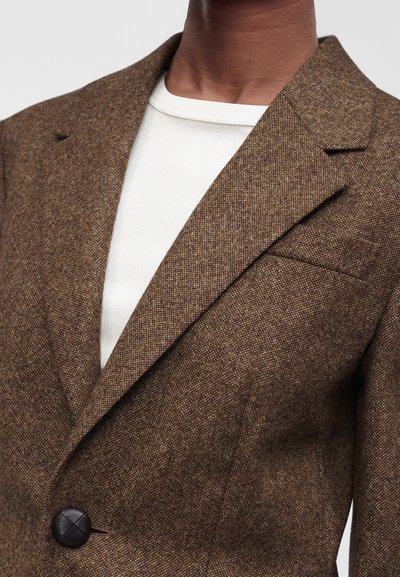 Blazer en laine marron texturé avec revers en encoche, porté sur un t-shirt blanc côtelé à col rond, boutonné sur le devant.