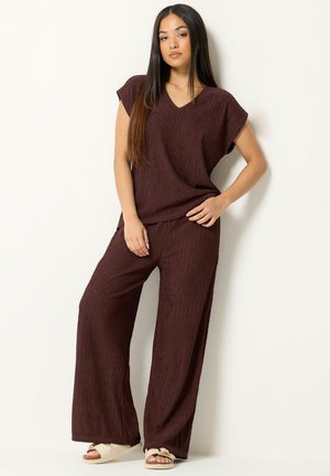 Femme debout contre un mur blanc portant un haut marron texturé à col en V et un pantalon large avec des sandales beiges.