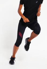Leggings atléticos negros de longitud a la rodilla, con paneles de malla en un tono más oscuro y un diseño ajustado. Complementados con zapatillas de correr negras.