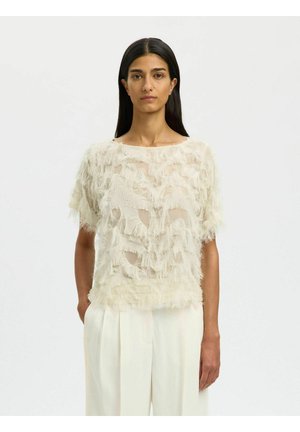 Frau mit langen dunklen Haaren trägt ein durchsichtiges cremefarbenes Kurzarm-Top mit strukturiertem Fransen-Design über weißen, hoch taillierten Hosen vor einem schlichten weißen Hintergrund.