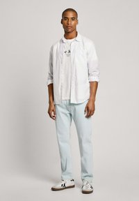 Camicia bianca a maniche lunghe sopra una maglietta bianca con testo nero, jeans azzurri chiari e sneakers bianche con dettagli neri. Sfondo neutro.