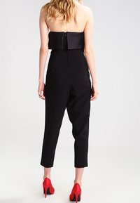 Vrouw in een strapless zwarte jumpsuit en rode hoge hakken, van achteren weergegeven tegen een effen lichte achtergrond.