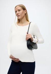 Έγκυος γυναίκα με λευκό μακρυμάνικο τοπ και σκούρο τζιν, κρατώντας την κοιλιά της με το ένα χέρι και μια μαύρη τσάντα στον ώμο της.