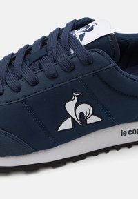 Mörkblå sneaker med en slät syntetisk ovandel, utrustad med en vit logotyp och en kontrasterande svart gummisula. Snörningen är i tyg och tungan är vadderad.