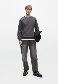 Szary sweter z okrągłym dekoltem, prążkowanymi mankietami i subtelnym logo. Noszony z wyblakłymi szarymi dżinsami i czarnymi butami, trzymając czarną czapkę.