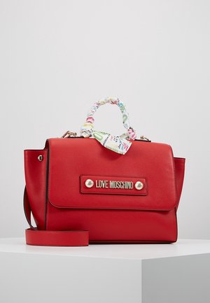 Punainen Love Moschino -käsilaukku kultaisella merkkikilvellä, päällystetty kangaskahva ja irrotettava olkahihna valkoisella pinnalla.