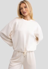 Sudadera oversized de color beige claro con hombros caídos, tejido texturizado y dobladillo con cordón. Combinada con pantalones beige a juego.