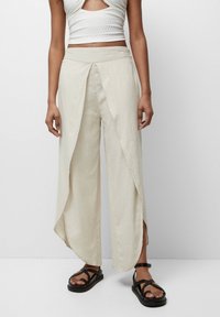 Pantalons larges en lin beige clair avec un design à l'avant enveloppant, des fentes sur les côtés et une texture lisse. Associés à des sandales à lanières noires.