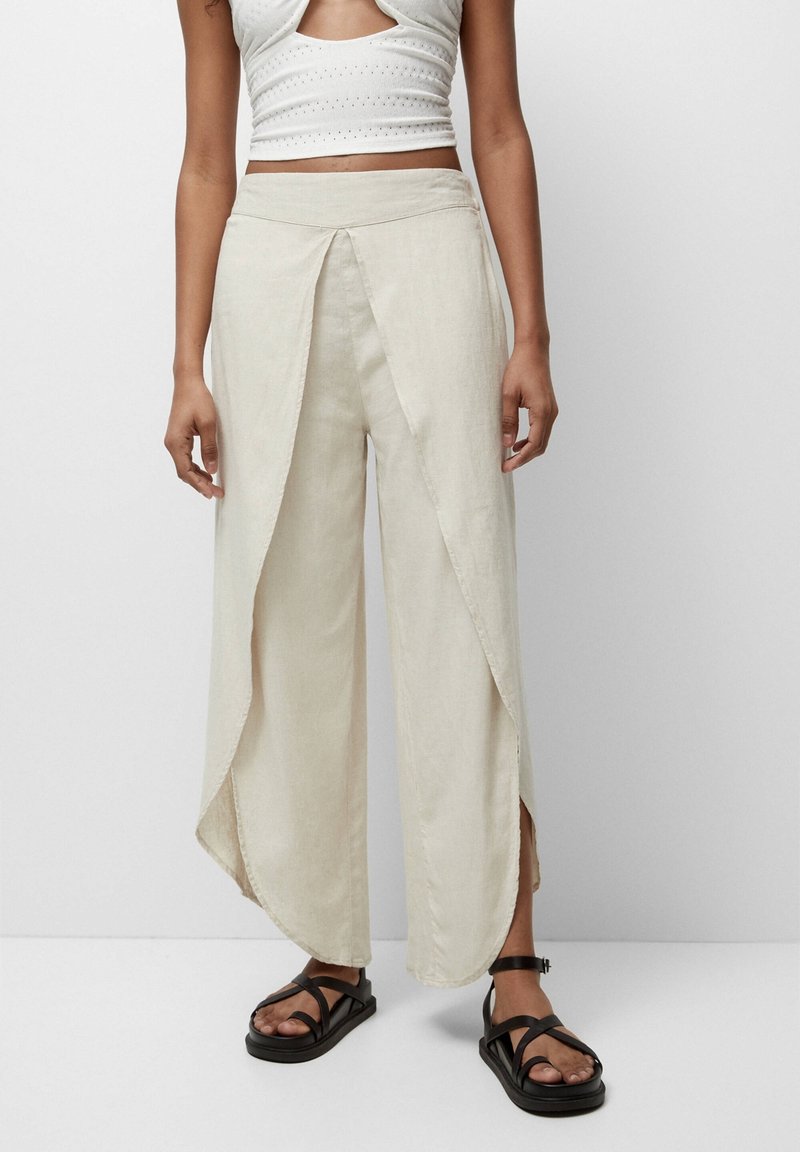 Pantalons larges en lin beige clair avec un design à l'avant enveloppant, des fentes sur les côtés et une texture lisse. Associés à des sandales à lanières noires.