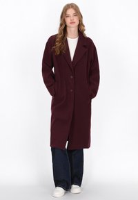Una giovane donna con capelli mossi indossa un lungo cappotto bordeaux scuro sopra una camicia bianca, jeans larghi blu e sneaker bianche, in piedi con le mani nelle tasche.
