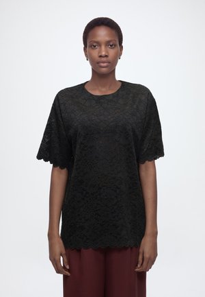 Kvinde stående, vendt fremad, iført en sort blonder kortærmet top og bordeaux bukser på en ensfarvet hvid baggrund.