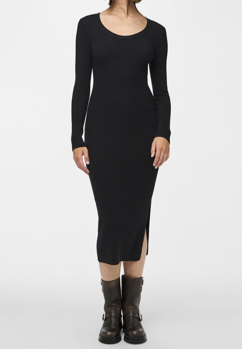 Femme portant une robe midi noire moulante à manches longues côtelée avec une fente latérale, associée à des bottes en cuir noires mi-mollet avec boucles.