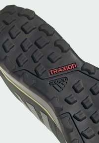Semelle de chaussure noire en caoutchouc avec des motifs de traction profonds, affichant "TRAXION" en rouge et "adidas" en noir ; bord arrondi et surface texturée.