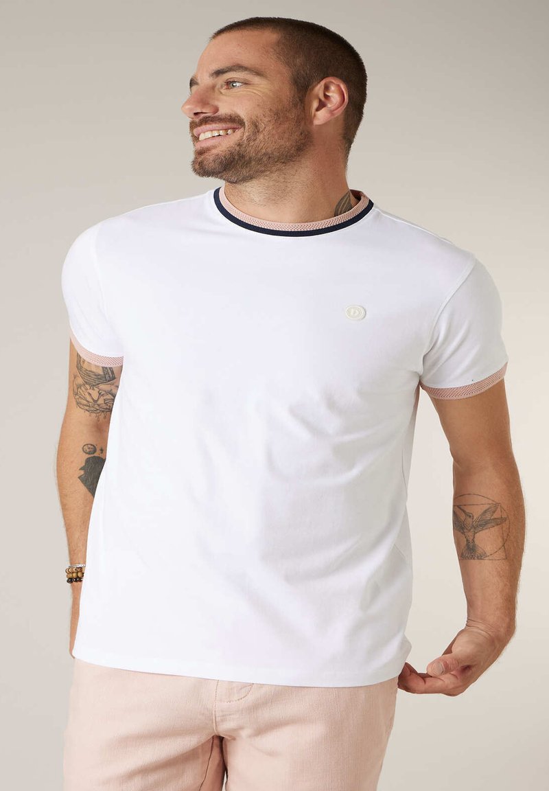 Homme souriant aux cheveux courts et tatouages, portant un t-shirt blanc avec des bordures roses et noires, associé à un pantalon rose clair sur un fond neutre.