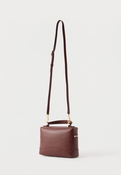 Καφέ δερμάτινο τσάντα χιαστί με ρυθμιζόμενο λουράκι, χρυσά εξαρτήματα και επάνω τσέπη με φερμουάρ, τοποθετημένη σε απλό ανοιχτό φόντο.