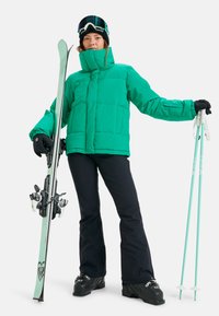Green insulated ski jacket, black ski pants, black gloves, holding teal ski poles and skis. Features goggles and a high collar design.